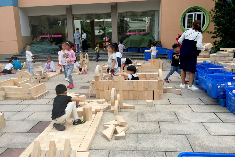 School children constructing a fortress and other forms out of wooden Anji Play blocks on a playground patio.